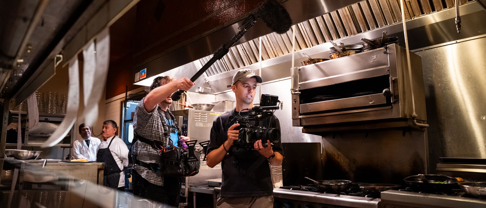 Tim Knoedler filming in a restaurant kitchen with a boom operator while the chefs look on